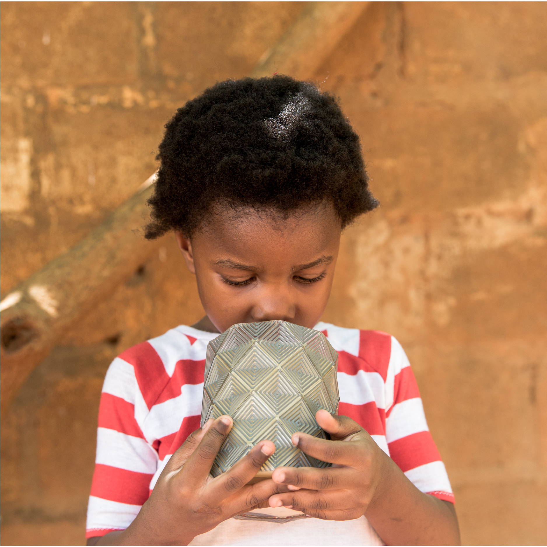 A young Zambian girl wearing a red and white striped shirt smells a Bridgewater Afternoon Retreat Candle. 