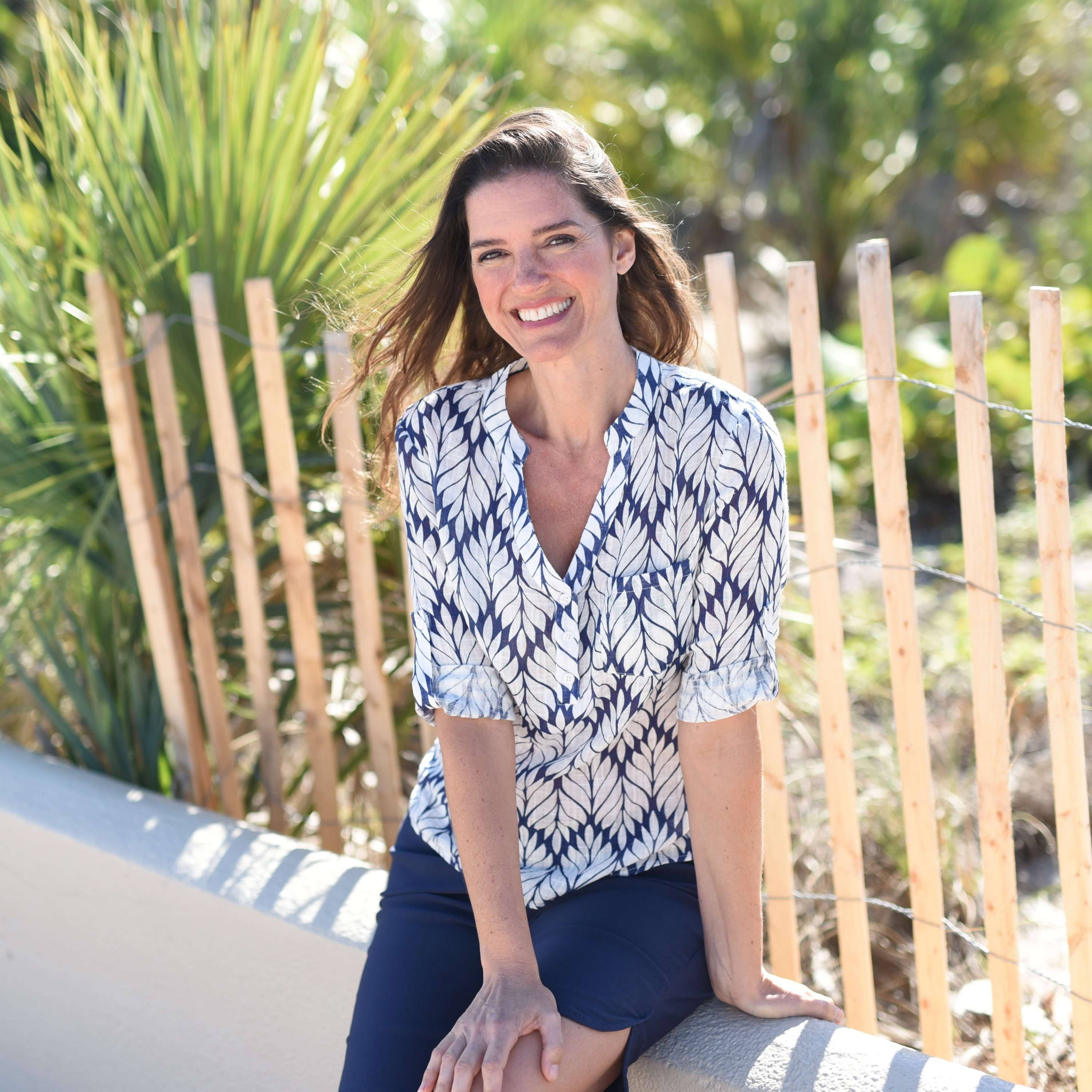 A brunette woman in a printed blouse and shorts smiles as she sits against a fence at the beach