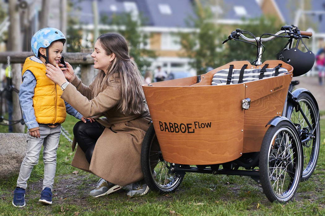 A mother fastens her child's helmet as they stand beside a Babboe Flow cargo bike.