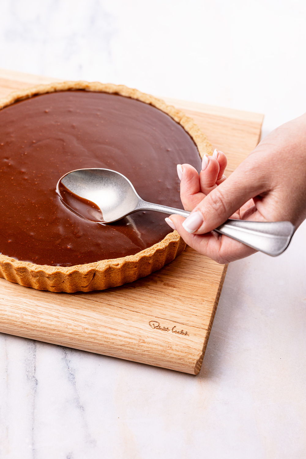 A hand using a spoon to serve chocolate tart on a wooden board.