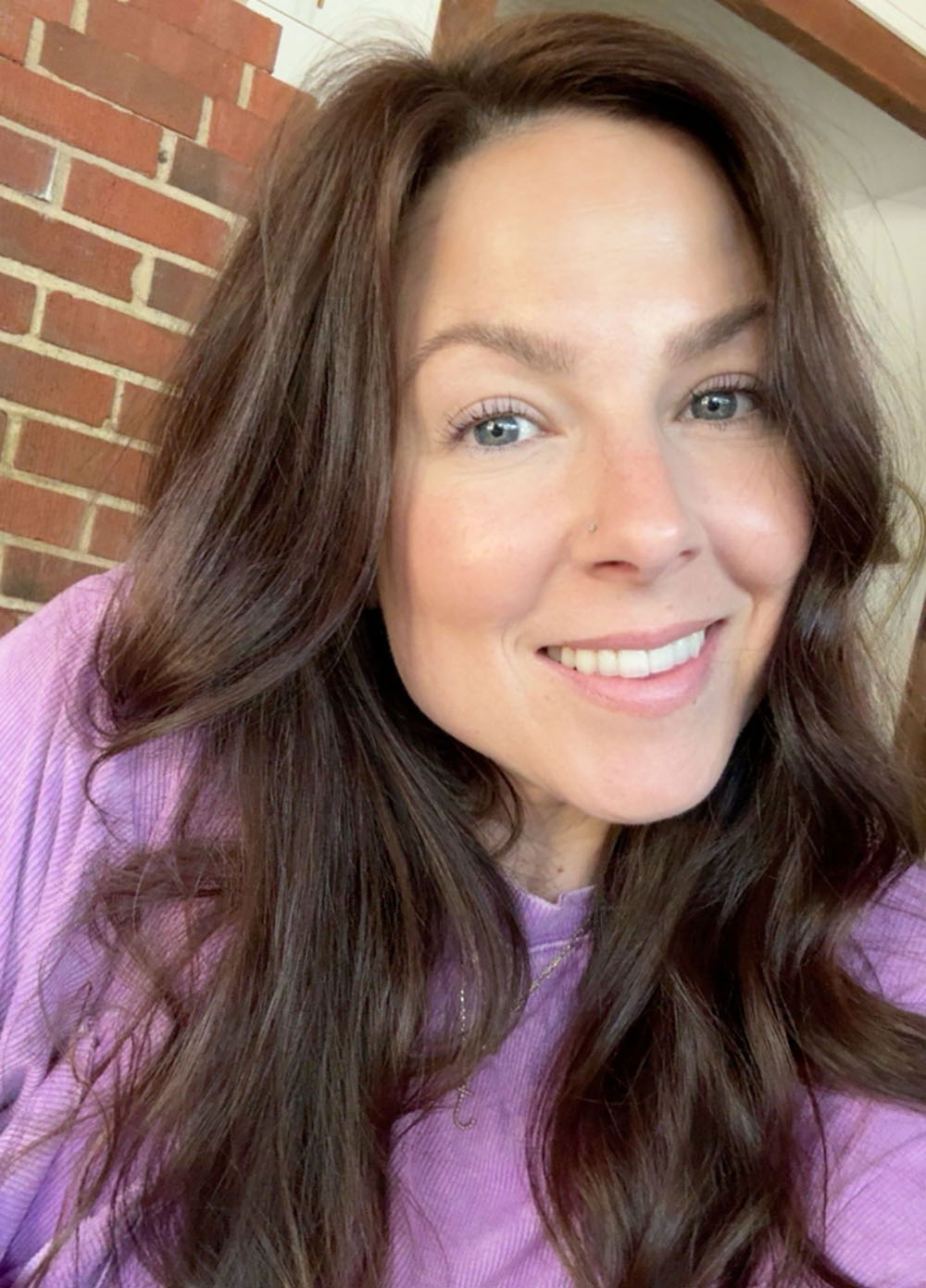 Smiling woman with long brown hair, wearing a purple shirt, against a brick wall.