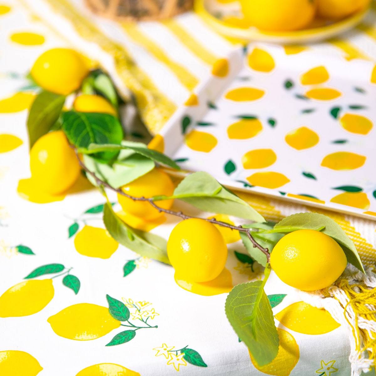 An artistic display of artificial lemons and green leaves artistically arranged on a lemon-print tablecloth. The bright yellow lemons and vibrant greenery are accentuated against a patterned backdrop, enhancing the summer-themed decor with a refreshing and lively visual appeal.