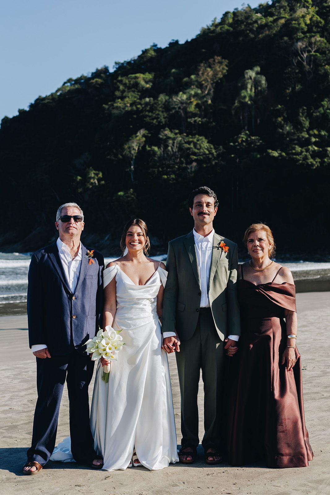 Four people in formal attire stand on a beach with a mountain backdrop.