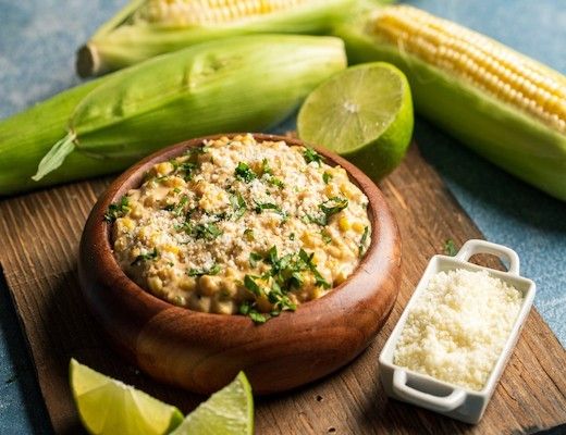 Bowl of corn dish with lime and cheese, surrounded by corn on a wooden board.