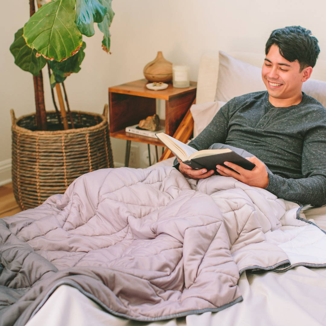 a man laying in bed reading a book while covered in the nest bedding luxury weighted blanket