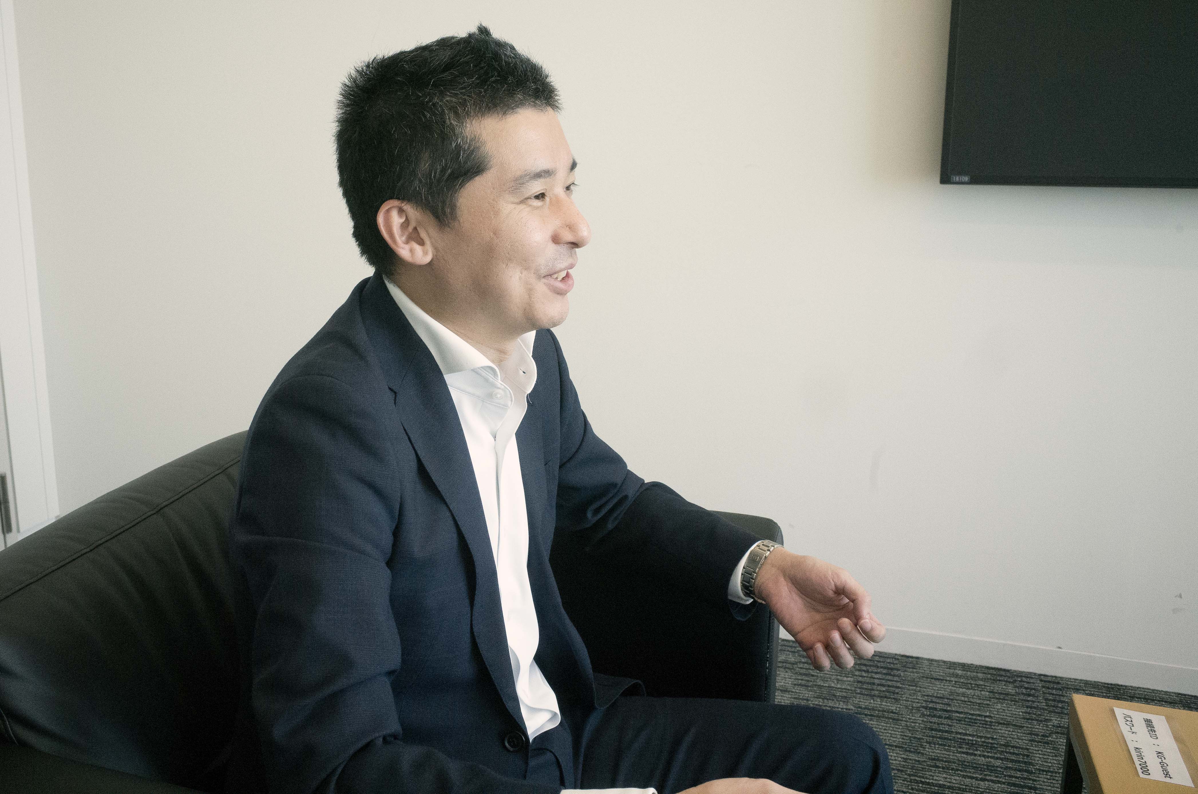 A man in a suit sitting on a couch, smiling.