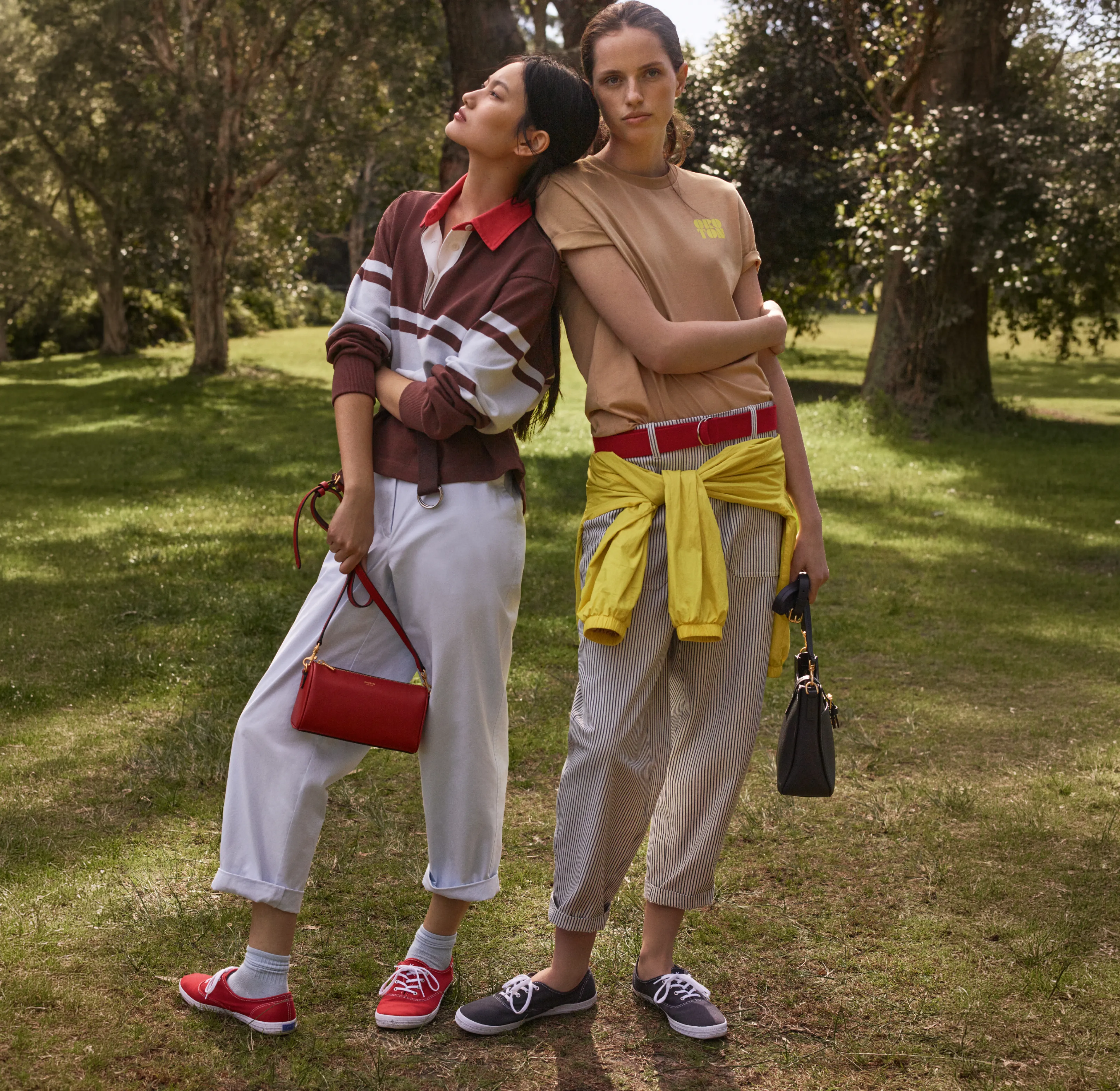 Two women posing outdoors in casual outfits, surrounded by trees.
