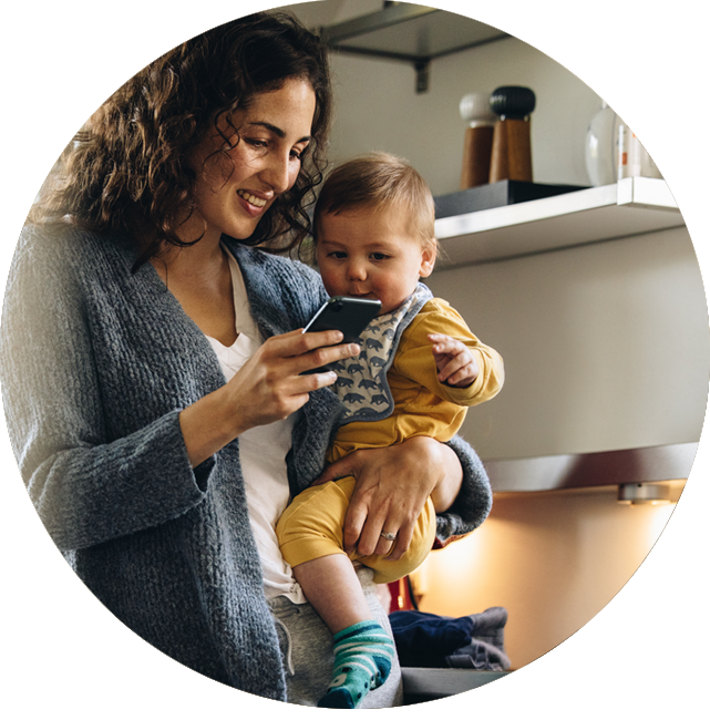 Woman holding a baby while looking at a smartphone in a kitchen.