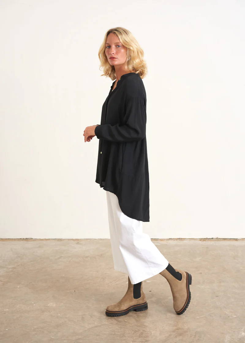 A model wearing an oversized black white with long sleeves and a dipped hem over white wide leg trousers and taupe, suede chelsea boots