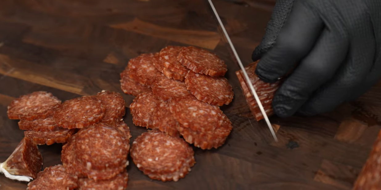 A hand in a black glove slicing pepperoni on a wooden board.