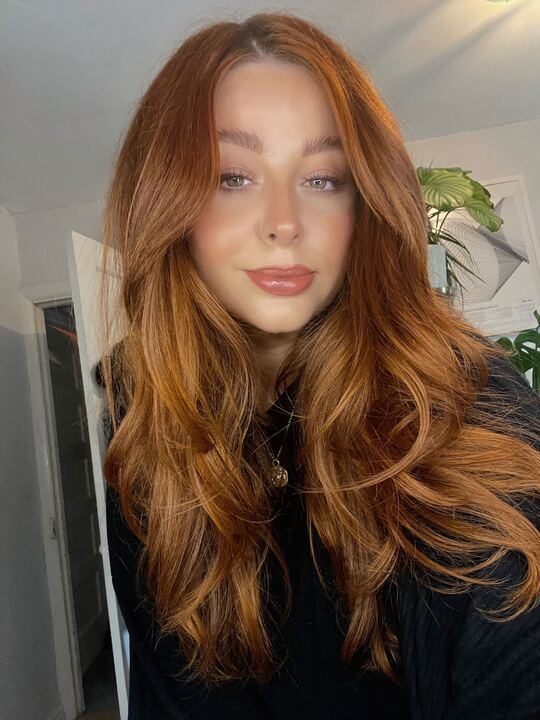 Young woman with long, wavy red hair, posing indoors with a plant.