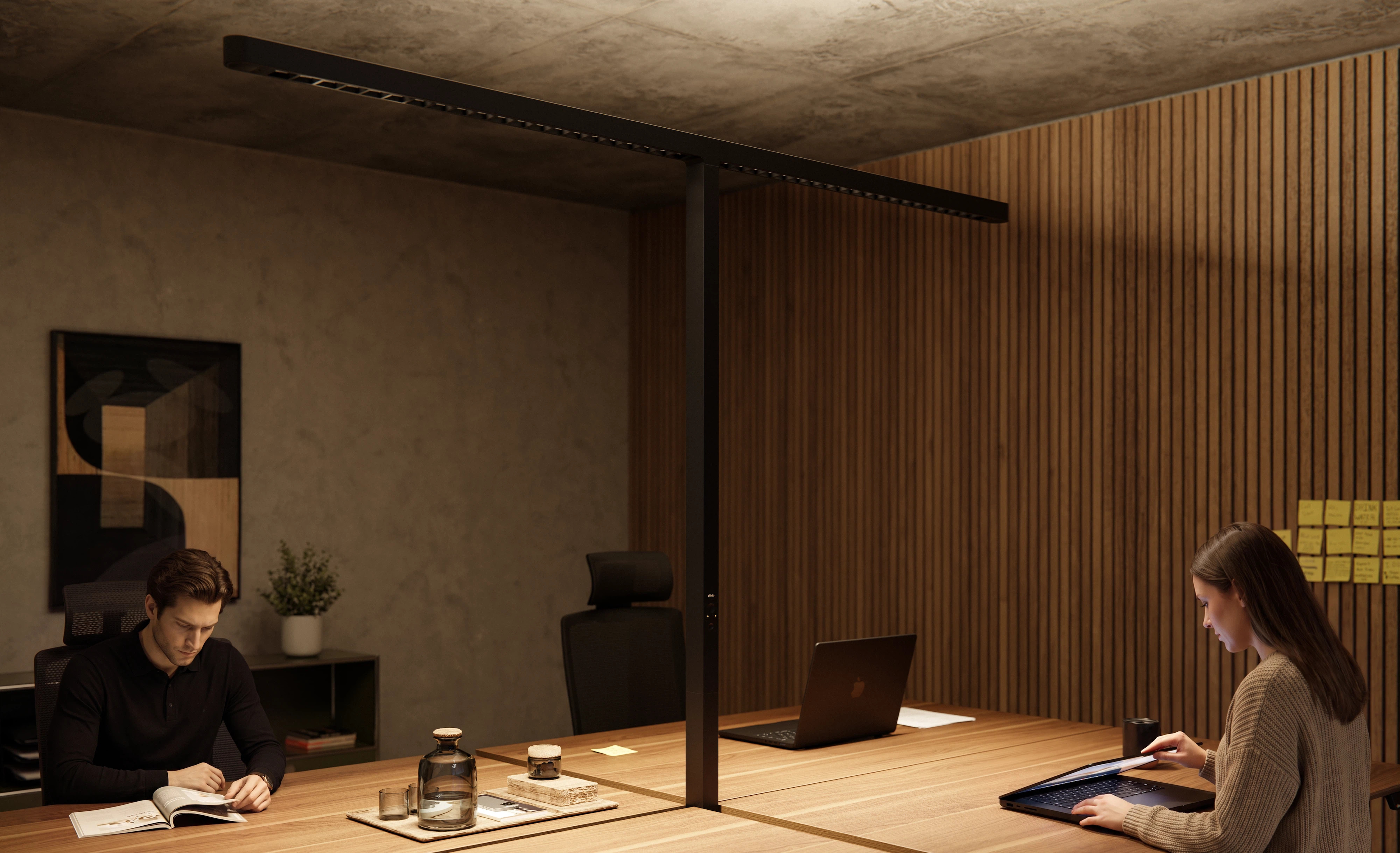 A man is reading on the left and a woman is working on a laptop on the right at a dual workstation, which is illuminated from the center by the black T-shaped floor lamp Lumino Pro Duo.