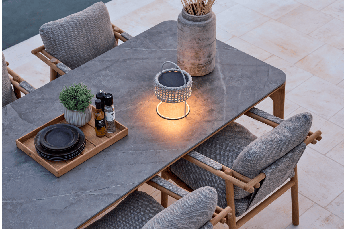 Evening view of a teak outdoor dining table with a gray stone-look top, cushioned dining chairs, and a glowing lantern centerpiece on a patio.