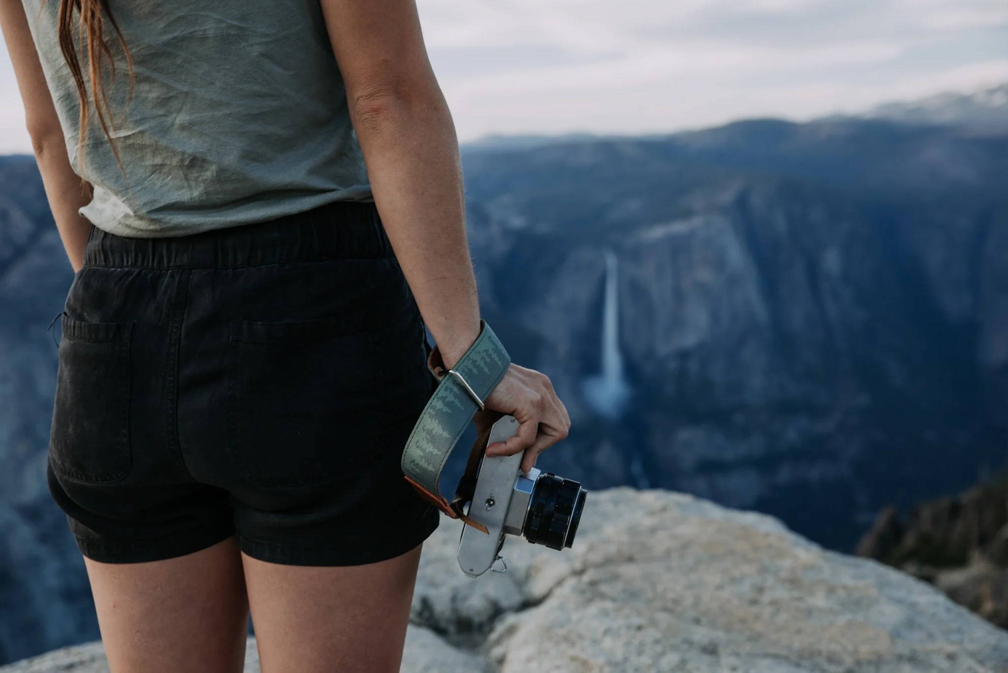 Woman standing on a rock edge with a camera and a camera strap and a blurred waterfall in the background. 