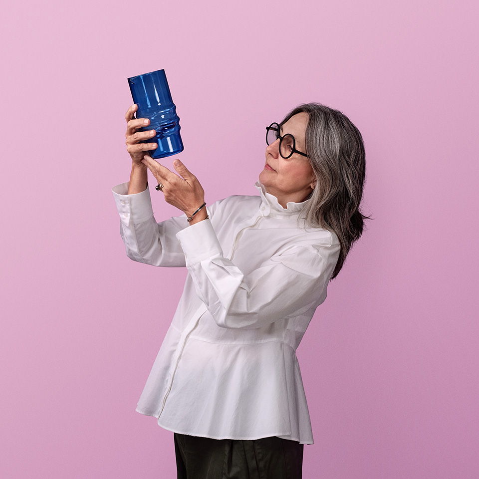 A woman in glasses examines a blue object against a pink background.