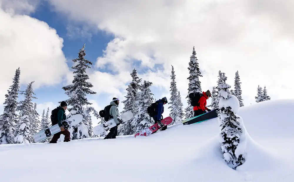 Group of snowboarders climbing a snowy hill among tall trees.