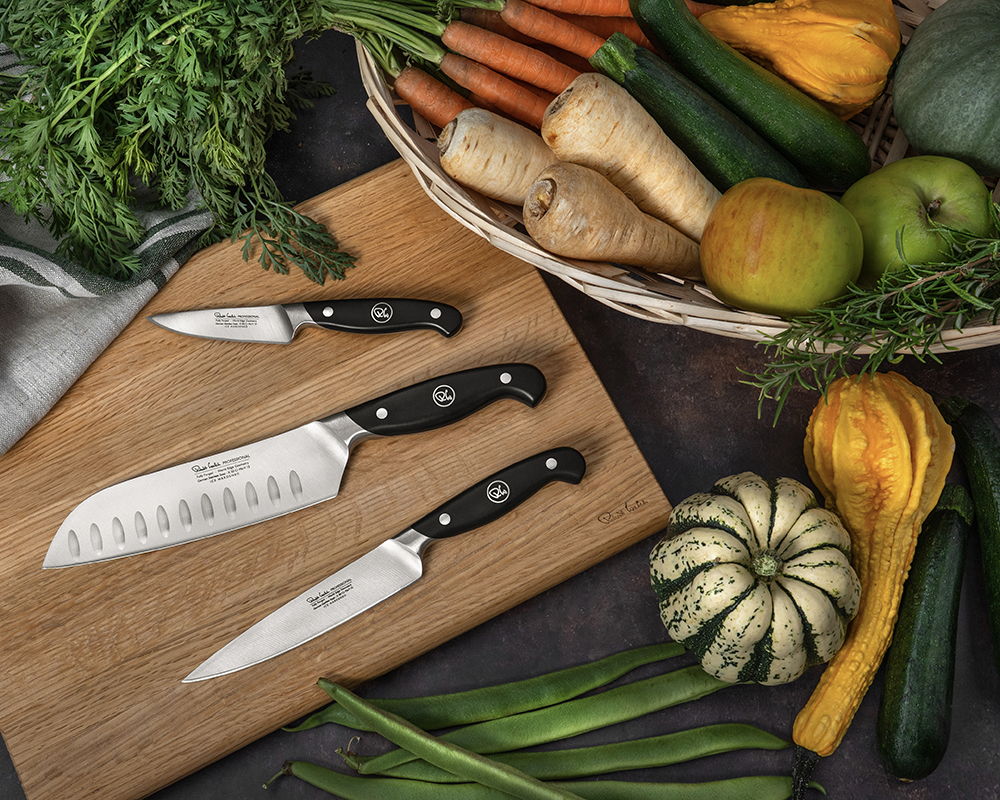 Three Professional Kitchen Knives on a chopping board with assorted vegetables in the background.