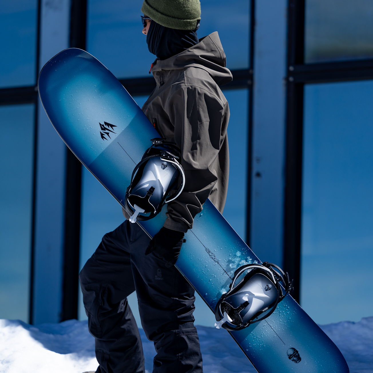 Person in winter attire holding a blue snowboard near a glass building.