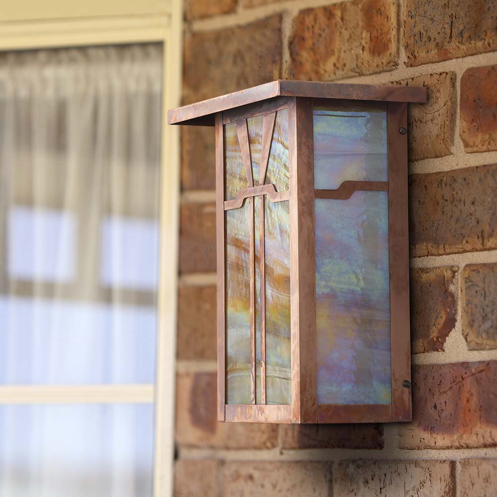 A linear, Craftsman style sconce with a raw copper finish installed against an exterior brick wall.
