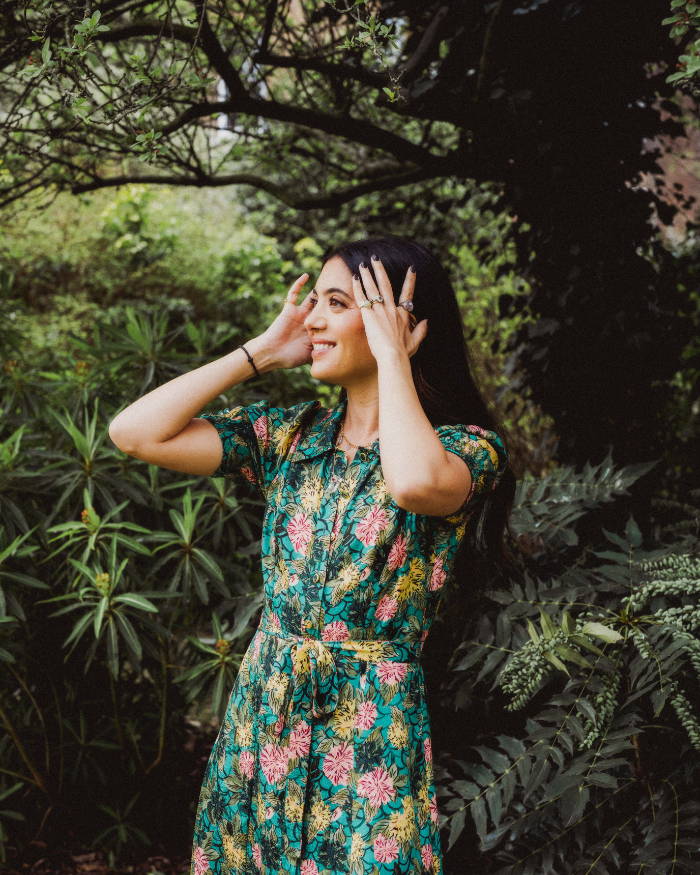Woman in a floral dress touching her hair, surrounded by greenery.