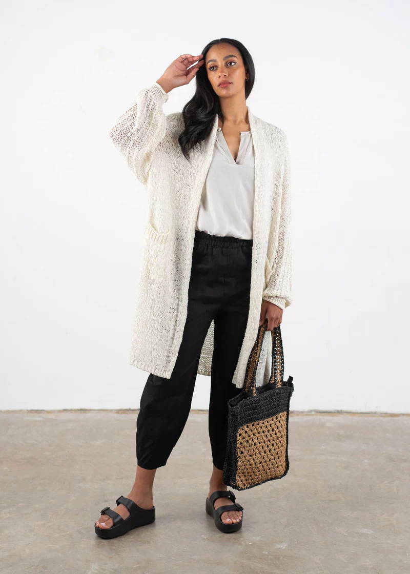 A model wearing a white long crochet cardigan over a off white top, black trousers, chunky platform slides and with a straw crochet bag