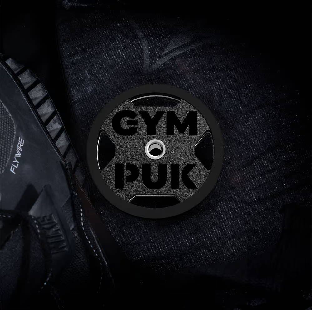 Black gym bag deodorizer shaped like a weight plate with 'GYMPUK' text, resting on black gym equipment