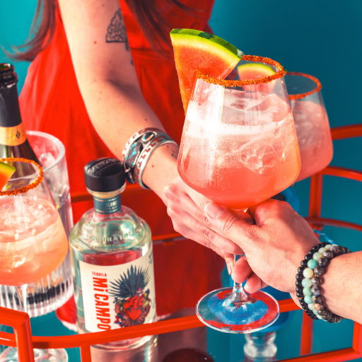 Two hands exchanging colorful watermelon cocktails on a vibrant bar cart.