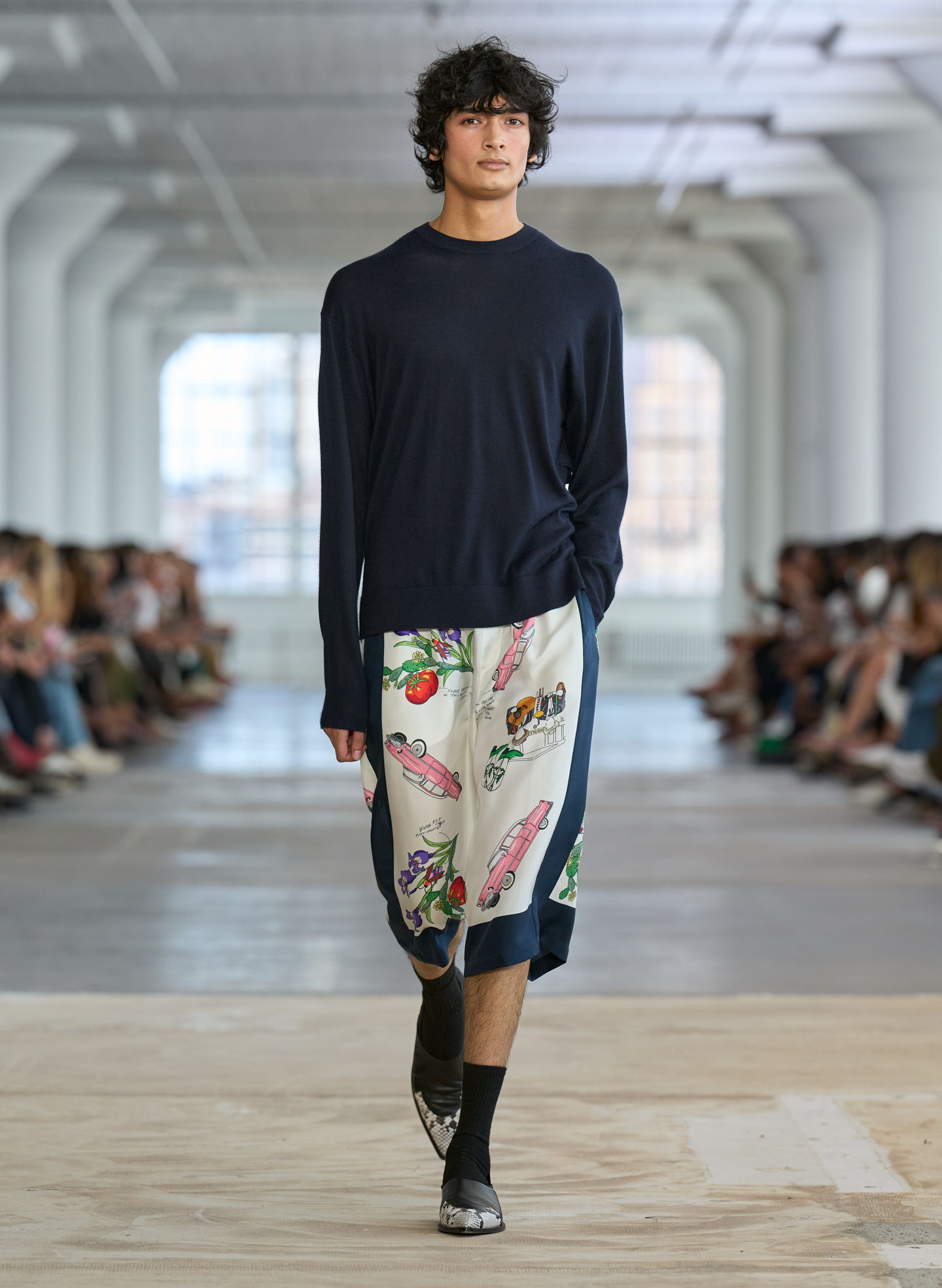 A model walks down the runway wearing a navy sweater and patterned skirt.