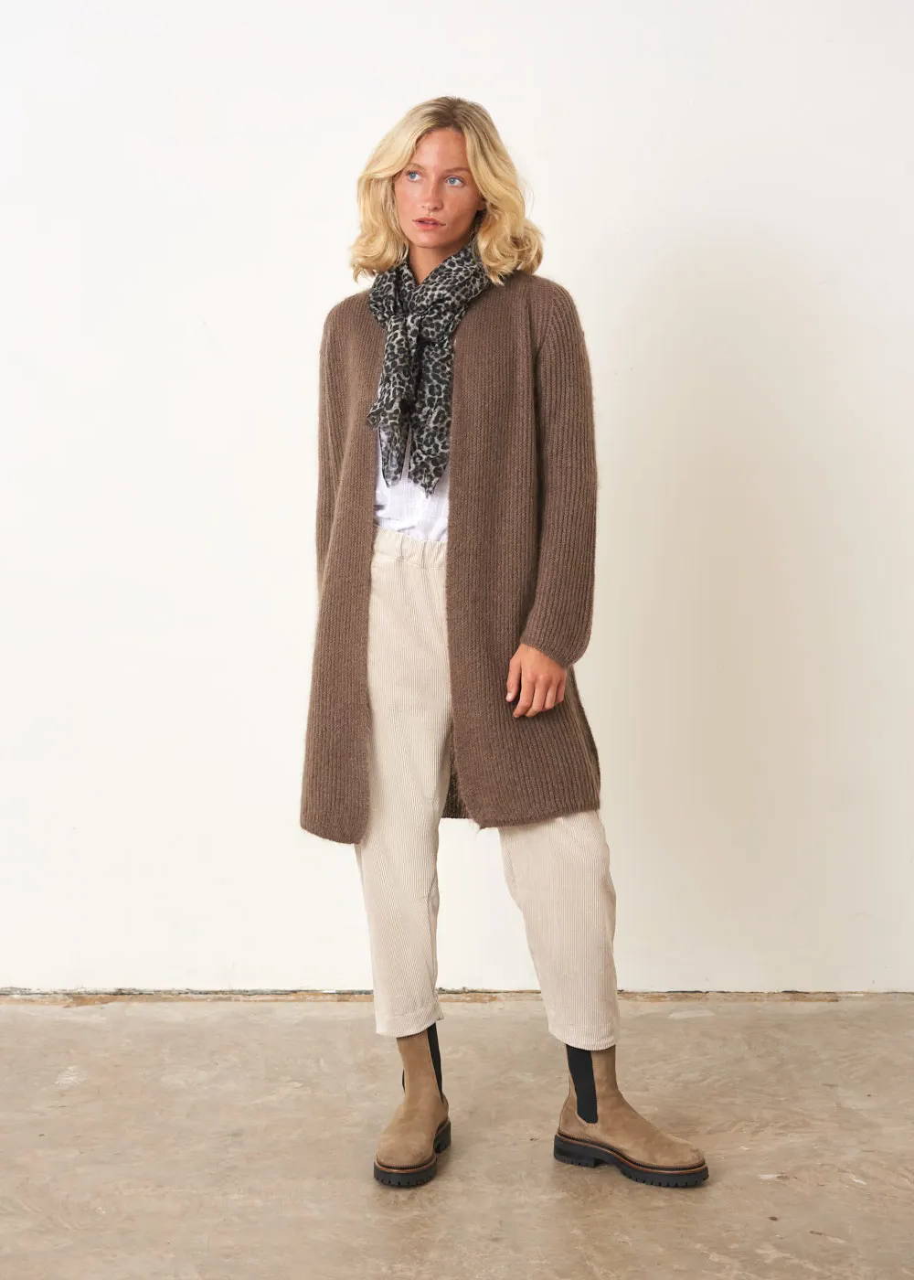 A model wearing a brown knitted midi cardigan over a white top, off white trousers with chunky taupe boots and a grey leopard print scarf