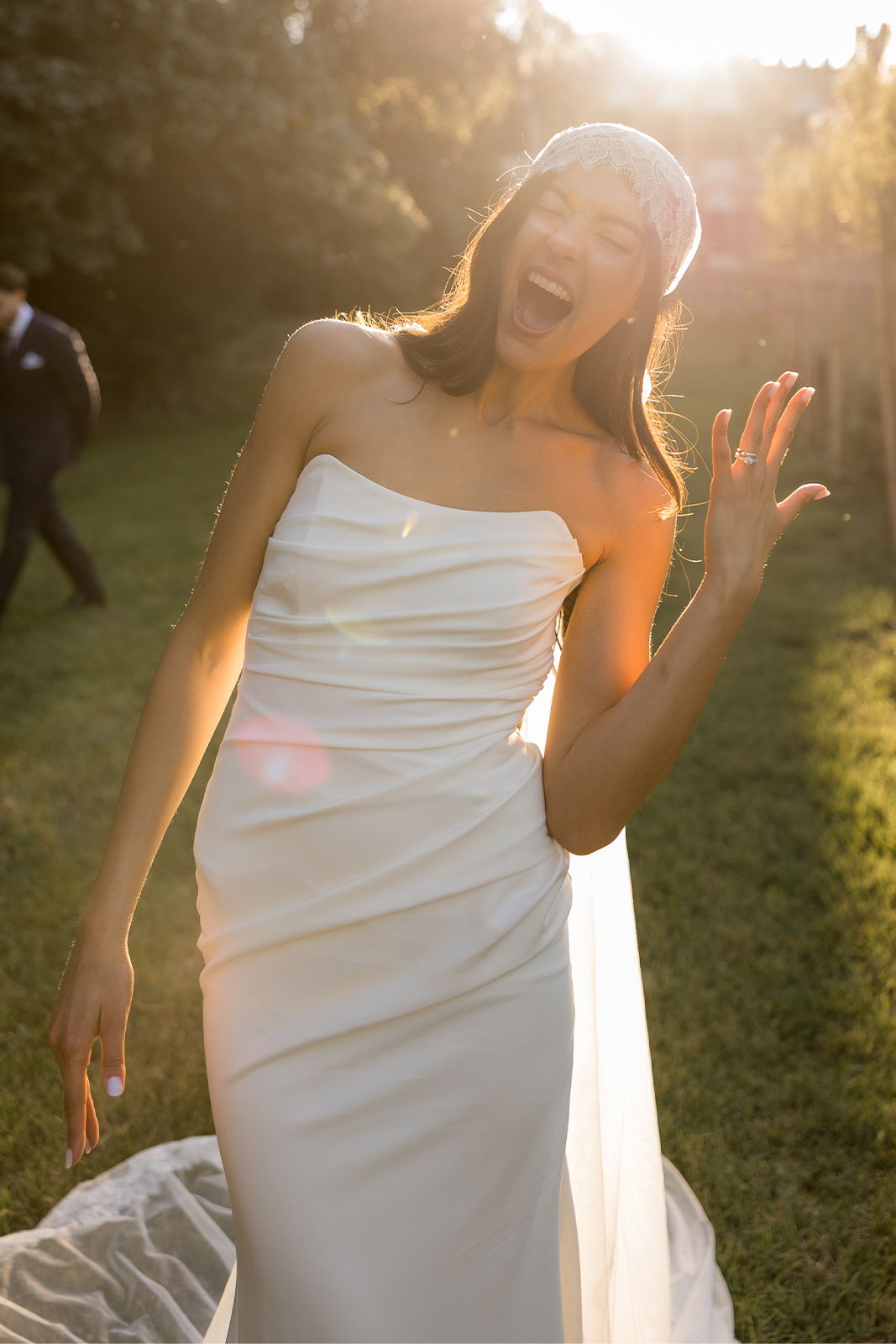 Smiling woman in a white dress waving in a sunlit outdoor setting.