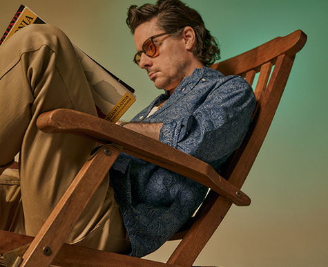 Person reading in a wooden chair, wearing sunglasses and a blue shirt.