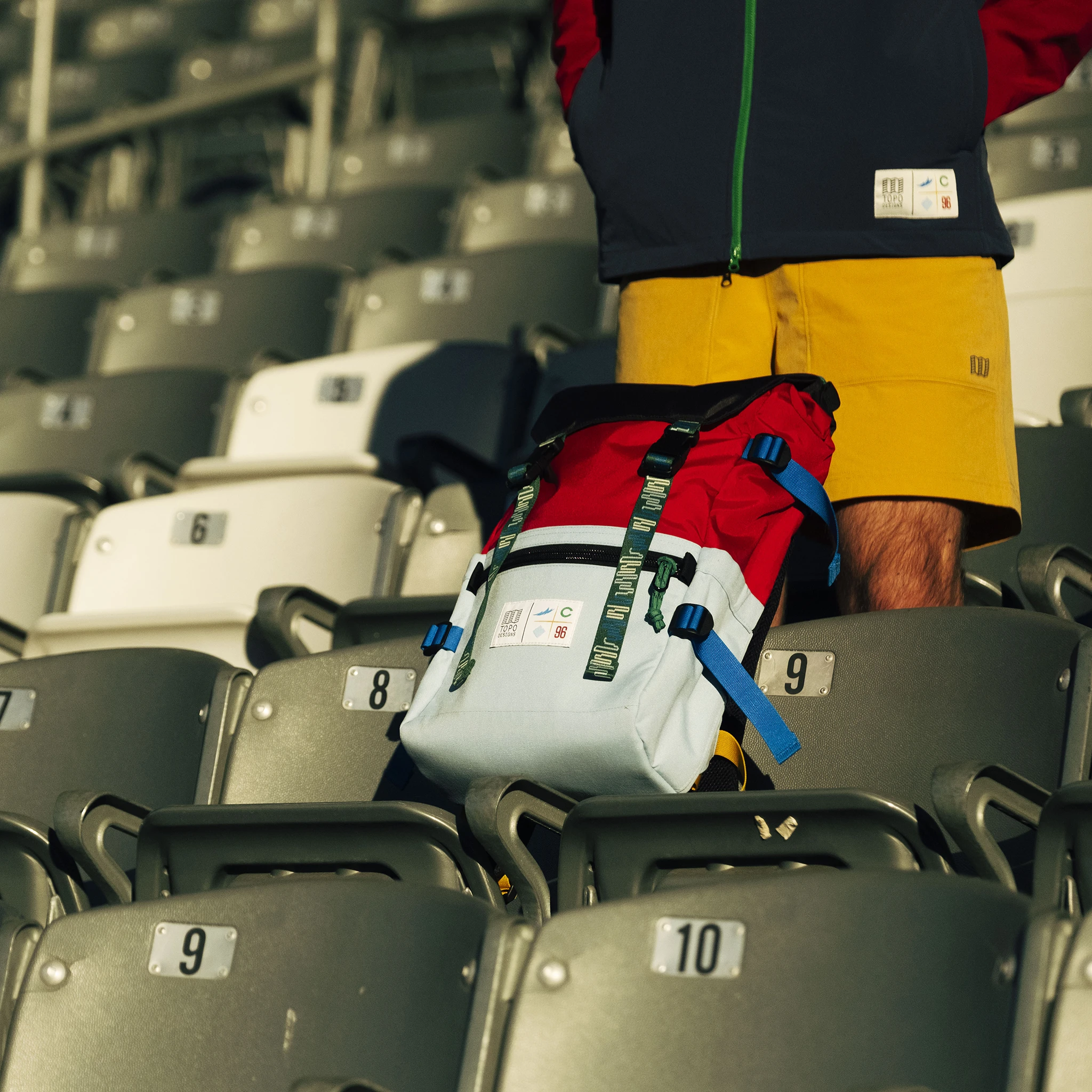 Person standing beside a colorful backpack on empty stadium seats.
