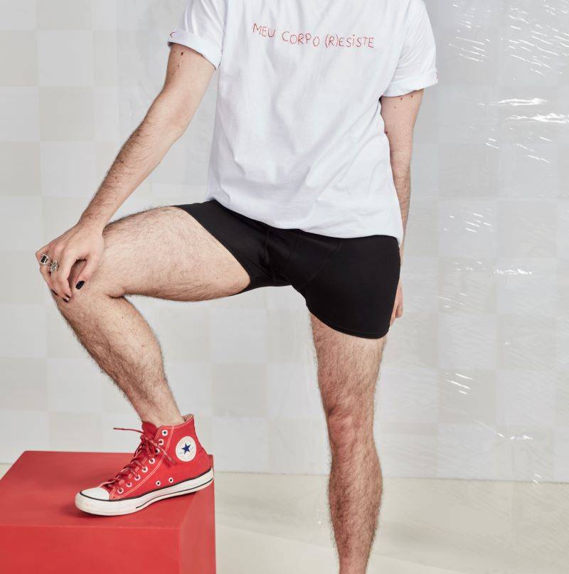 Foto de tronco e pernas de homem usando camiseta escrito meu corpo resiste, usa cueca boxer absorvente e tênis vermelho de cano médio