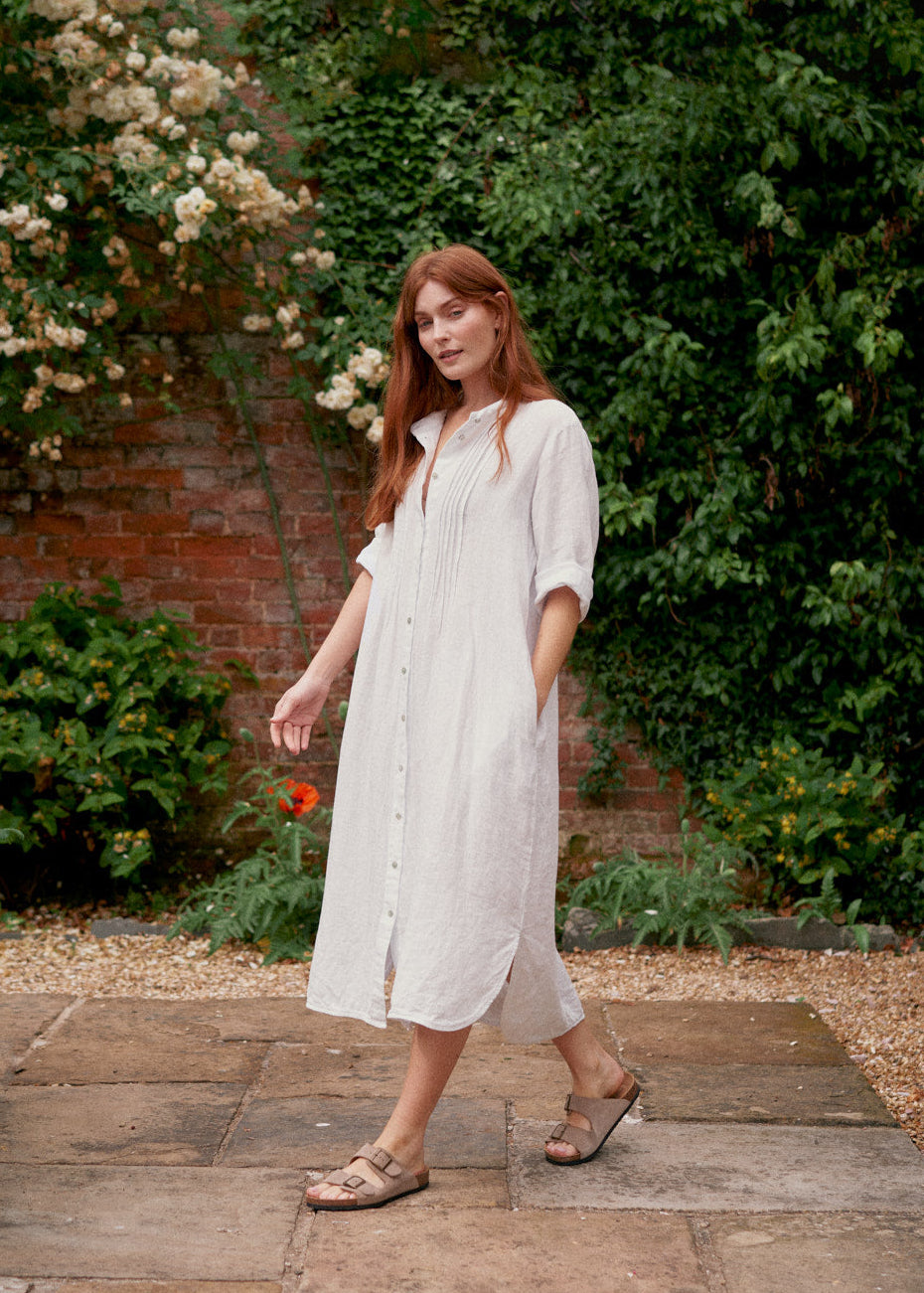 Woman wearing white linen shirt dress with button up detail and pleated from.