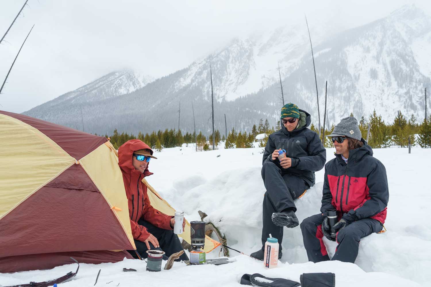 Three people sitting in snow near a tent, surrounded by mountains.