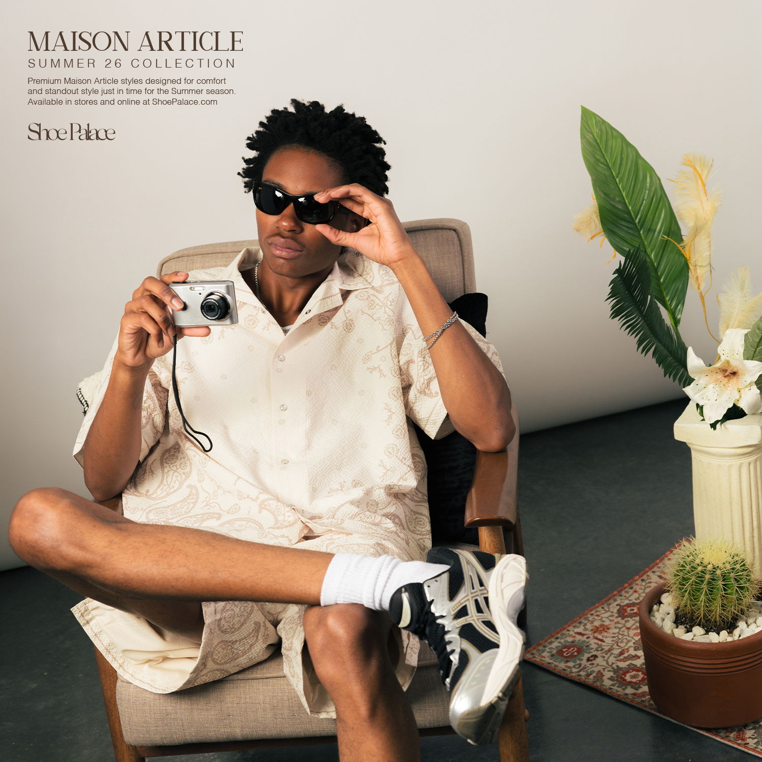 Person sitting with a camera, wearing sunglasses, in a stylish indoor setting.