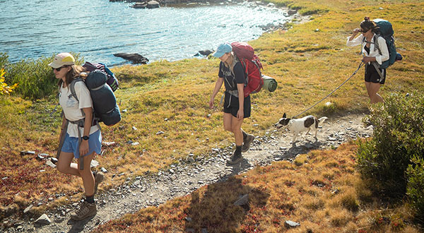 Three hikers with backpacks walking along a trail by a lake.