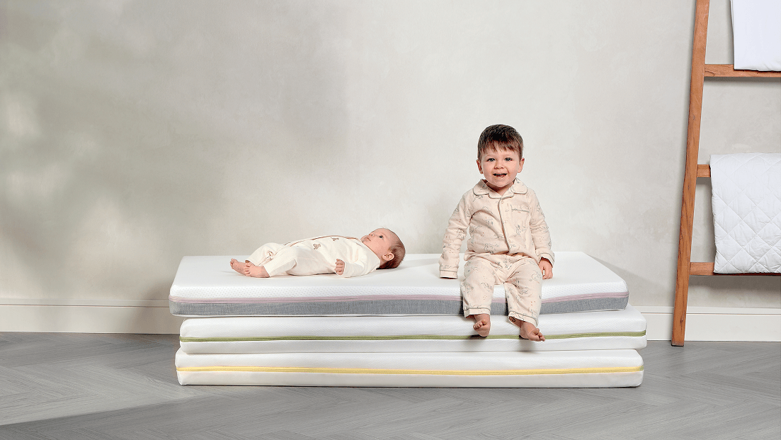 A toddler sitting on a mattress next to a sleeping baby.