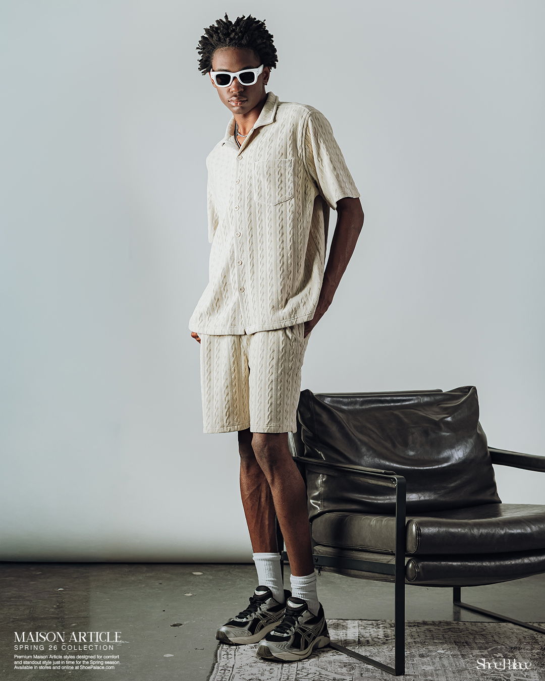 A person in light clothing stands next to a chair, wearing sunglasses and sneakers.
