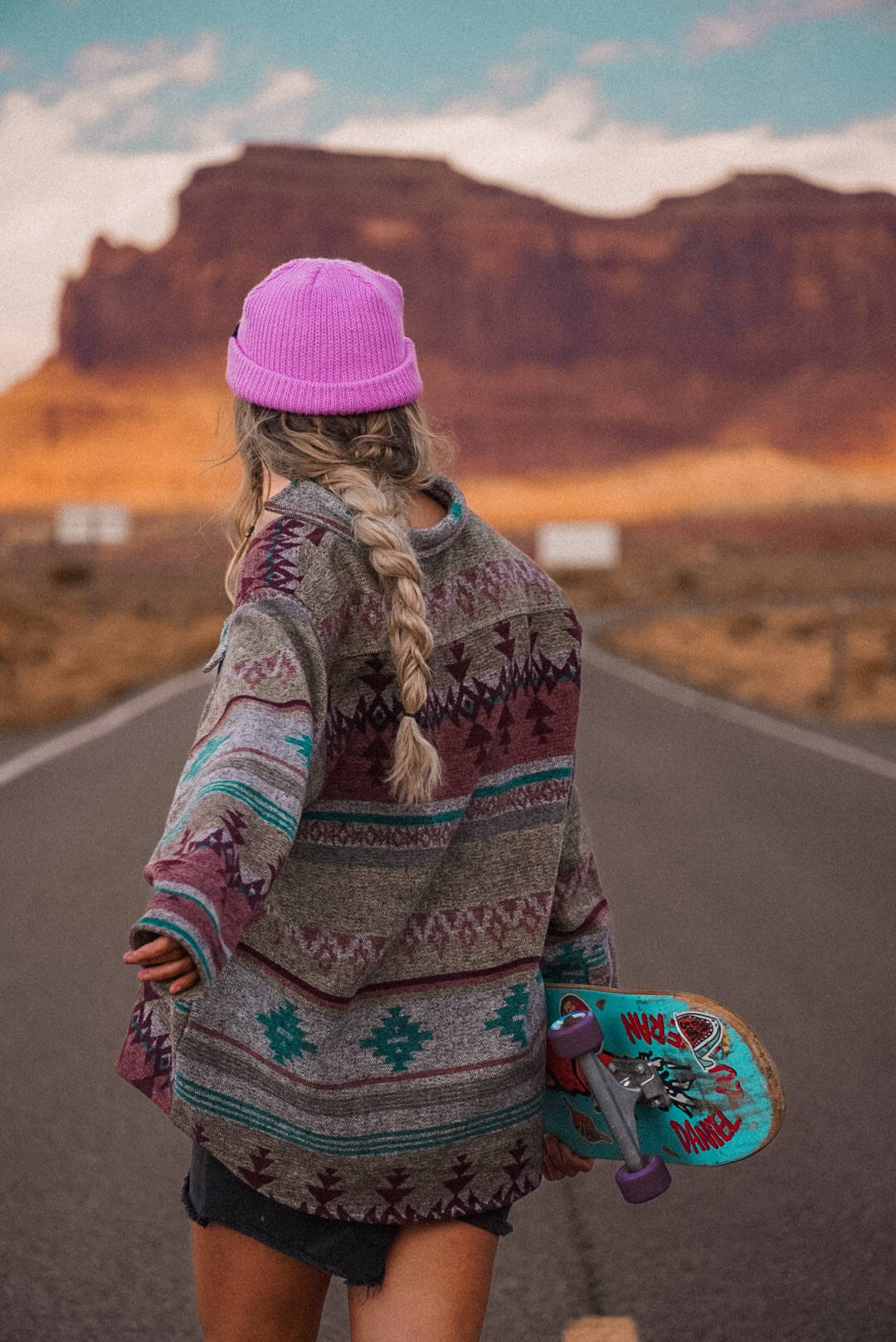 A woman holding her skateboard on an empty road, wearing a bright pink beanie in Utah