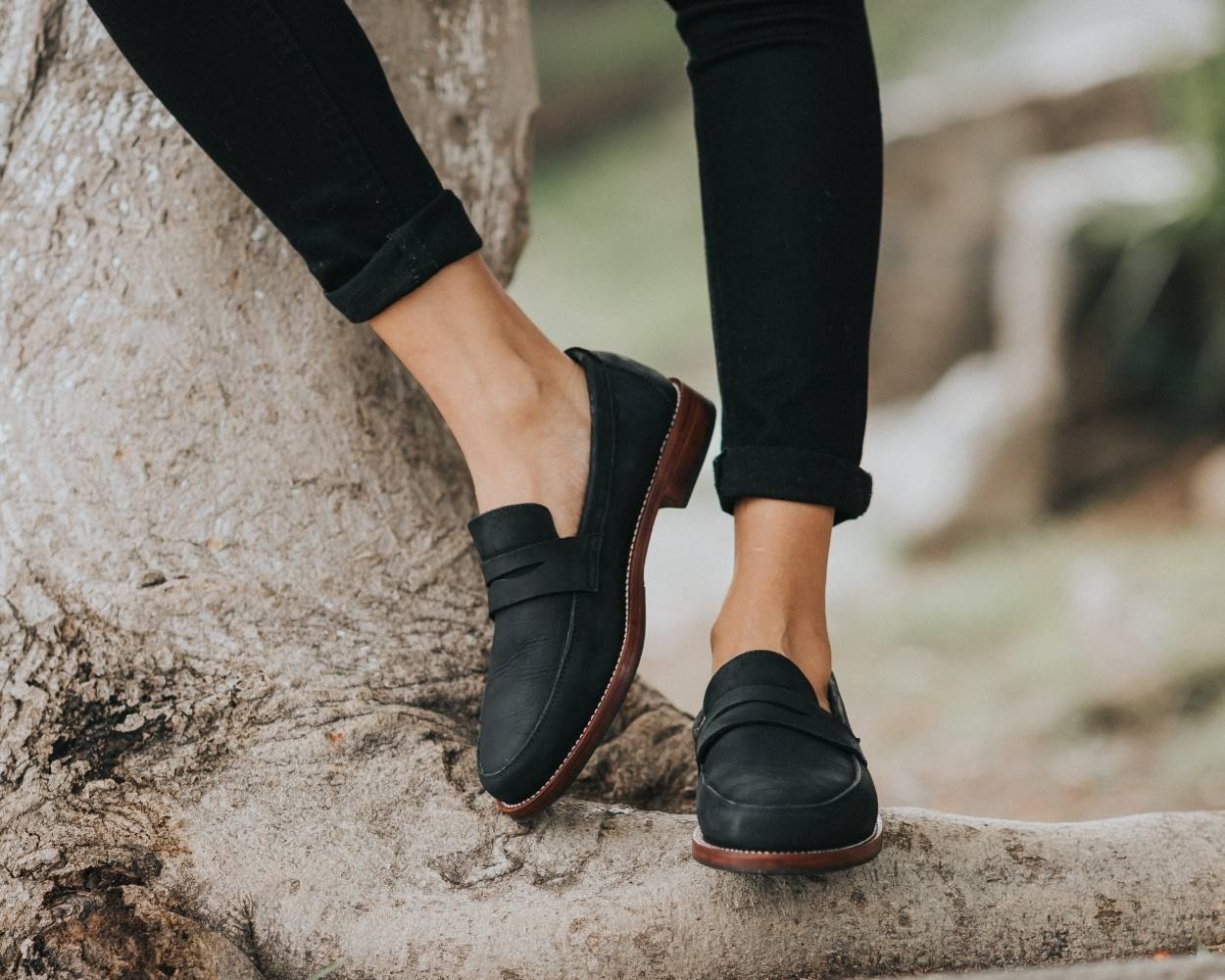 A woman standing on a large tree trunk, showcasing the handcrafted Adelante Sofia women's wide width leather loafers in classic black.