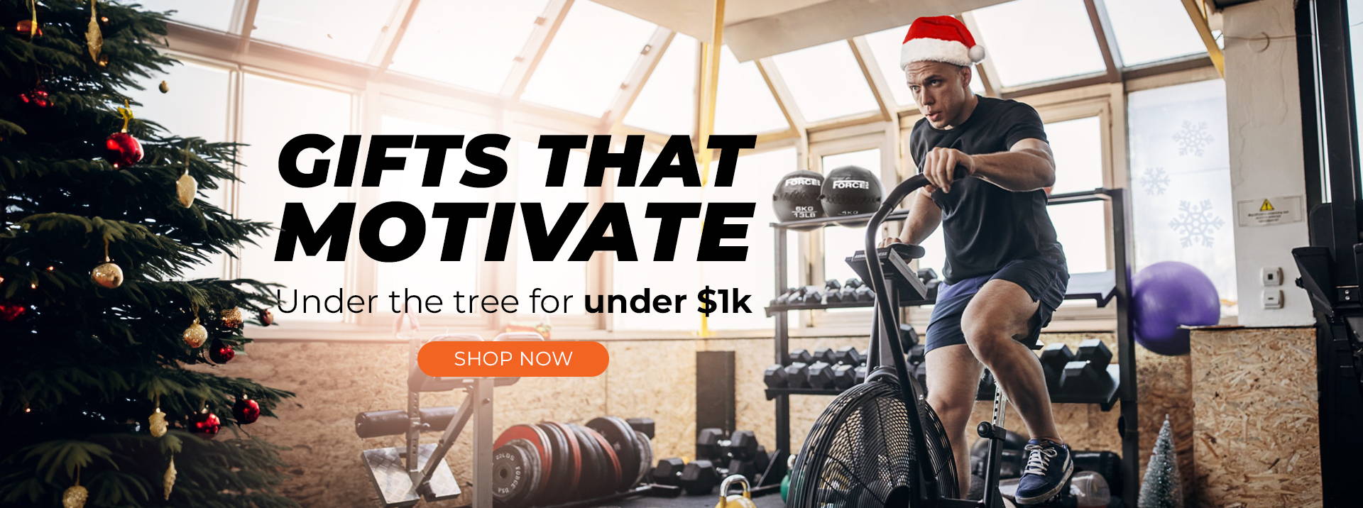 Man exercising on an Air Bike in a home gym, surrounded by gym equipment and festive Christmas decorations, with text overlay: 'Gifts that Motivate - Find the Perfect Fitness Present Under Your Tree for Under $1k.