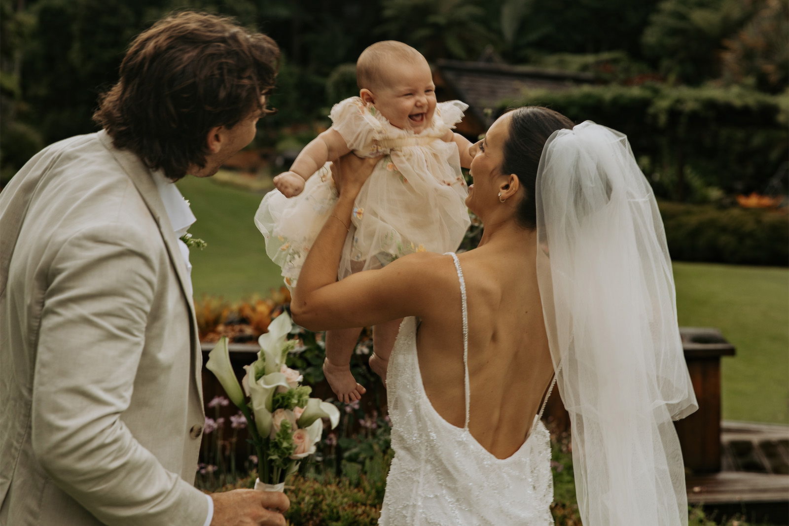 A couple joyfully holds their baby during a wedding celebration outdoors.