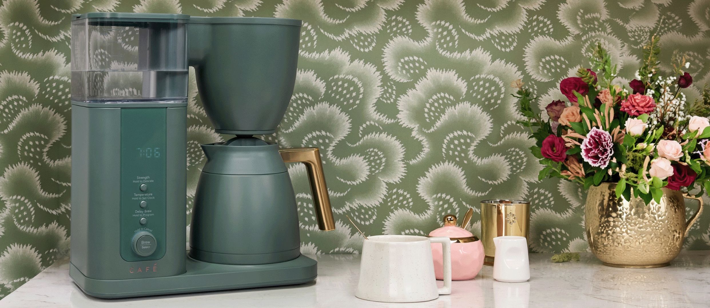 Cafe Coffee maker beside flowers and decorative items on a countertop.