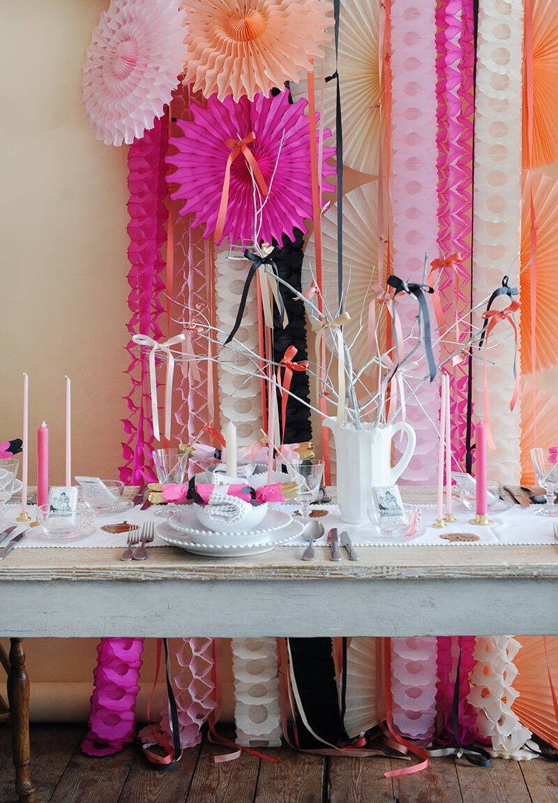 A table laid for a hen do with white pearl china, pink candles and a paper party decoration backdrop of fans and garlands in pinks, cream and black.