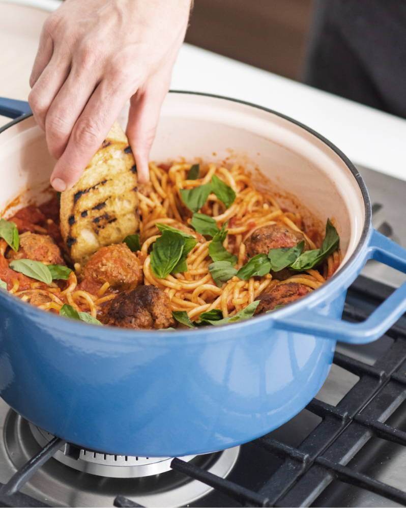  A blue Misen Dutch Oven filled with spaghetti and meatballs, with a hand dipping toasted bread into the red sauce.