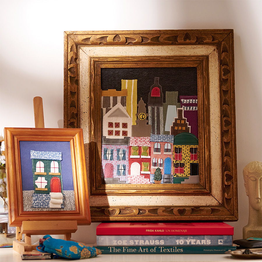 Two framed paintings of colorful buildings on a shelf with books.