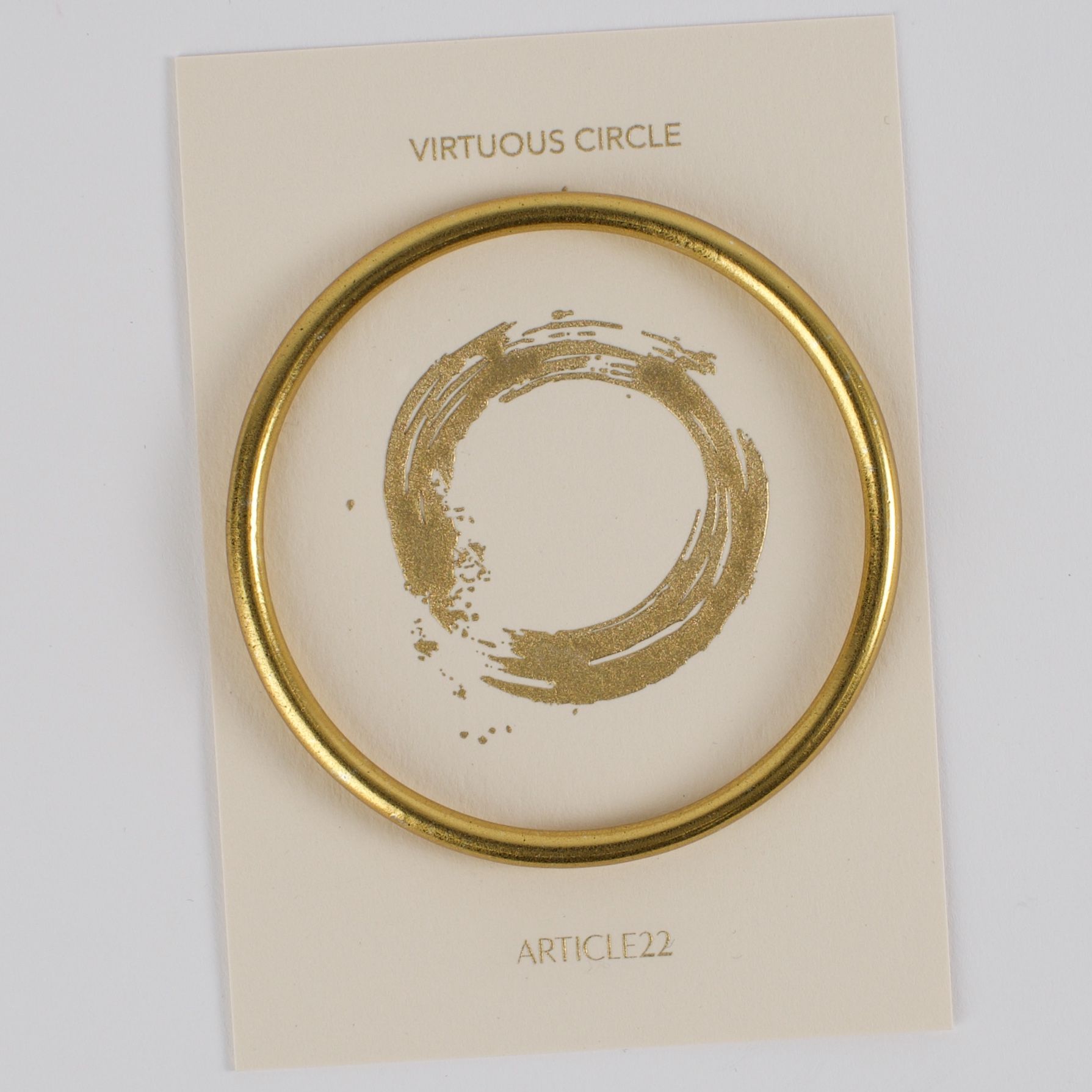 A gold bangle bracelet on a card with a circular brushstrokes symbolizing virtue.