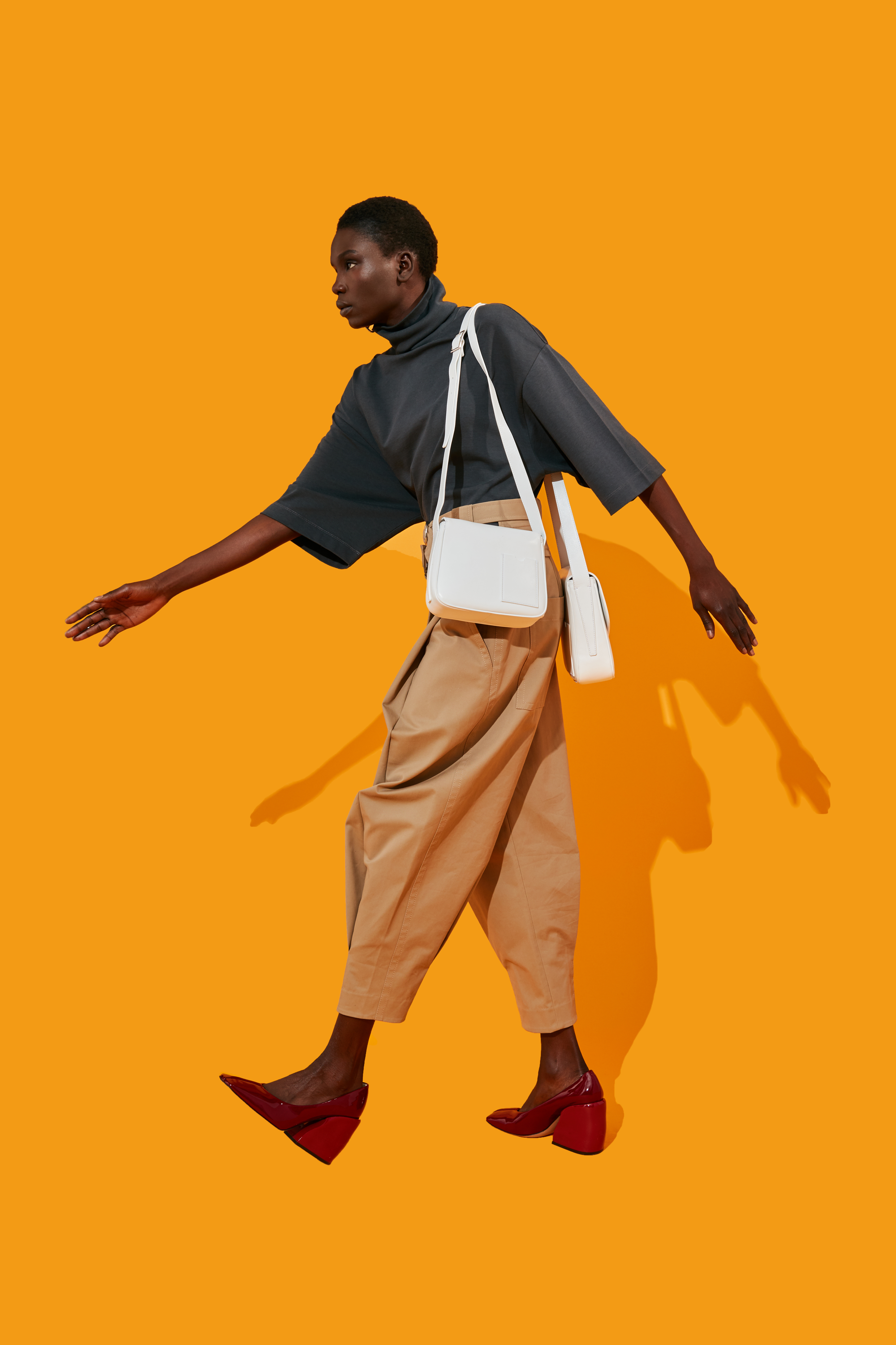 A person walking against a bright orange background, wearing stylish clothing and accessories.