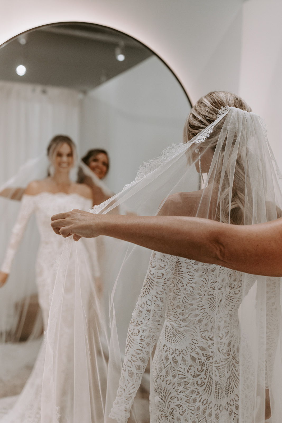 Bride adjusting her veil while admiring her dress in a mirror.